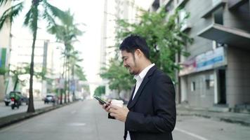 un hombre de negocios asiático sonriendo con un traje negro camina por las calles de la ciudad para trabajar. en la mano sosteniendo una taza de café usando un teléfono inteligente. concepto de estilo de vida urbano. video