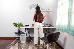 Attractive young beautiful asian woman working with laptop and document while sitting at the indoors living room office as a freelancer, e-coaching working, remotely or work from home concept.