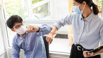 Two Asian colleagues in medical masks avoid a handshake when meeting in the office greeting with bumping elbows during coronavirus COVID-19 epidemic in office, Social distancing concept.