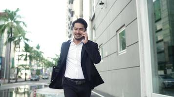 empresário asiático sorrindo vestindo um terno preto é correr nas ruas da cidade para trabalhar. na mão segurando um laptop usando smartphone. conceito de estilo de vida urbano.
