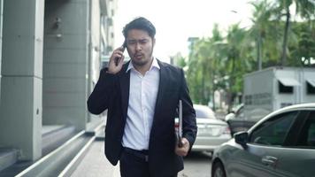 un hombre de negocios asiático sonriendo con un traje negro se apresura a correr por las calles de la ciudad para trabajar. en la mano sosteniendo una computadora portátil usando un teléfono inteligente. concepto de estilo de vida urbano. video