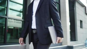 segue la visualizzazione dei caratteri. uomo d'affari che indossa un abito nero sta camminando per le strade della città per andare a lavorare. in mano che tiene un computer portatile. concetto di stile di vita urbano. video