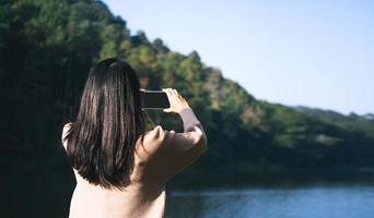 ella se relaja en el lago en el bosque en la montaña usa el teléfono inteligente para la red social. foto