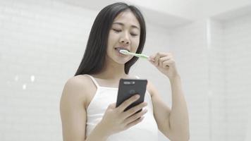mulher escovando os dentes e lendo a mensagem no telefone do banheiro. menina com smartphone usando escova de dentes, verificando redes sociais. video