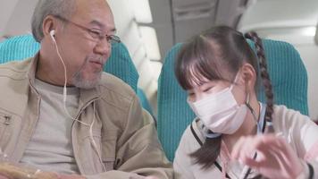 Happy Asian grandfather talking and laughing with granddaughter at seat in airplane cabin. family trip travel vacation. little girl wearing protective mask video
