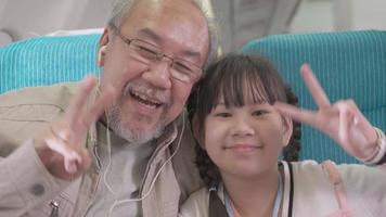 Happy Asian grandfather and granddaughter showing peace sign finger and mini heart looking at camera at seat in airplane cabin. family trip travel vacation video