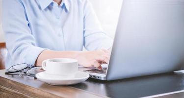 Business concept. Woman in blue shirt typing on computer with coffee on office table, backlighting, sun glare effect, close up, side view, copy space photo