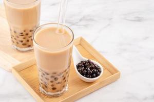 Tapioca pearl ball bubble milk tea, popular Taiwan drink, in drinking glass with straw on marble white table and wooden tray, close up, copy space. photo