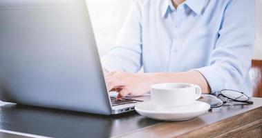 Business concept. Woman in blue shirt typing on computer with coffee on office table, backlighting, sun glare effect, close up, side view, copy space photo