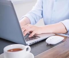 Business concept. Woman in blue shirt typing on computer with coffee on office table, backlighting, sun glare effect, close up, side view, copy space photo