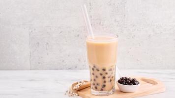 Tapioca pearl ball bubble milk tea, popular Taiwan drink, in drinking glass with straw on marble white table and wooden tray, close up, copy space. photo