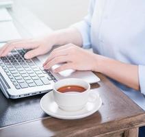 Business concept. Woman in blue shirt typing on computer with coffee on office table, backlighting, sun glare effect, close up, side view, copy space photo