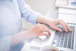 Business concept. Woman in blue shirt typing on computer with coffee on office table, backlighting, sun glare effect, close up, side view, copy space photo