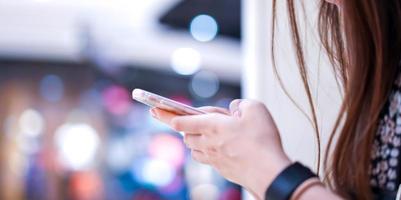 Young girl is holding smart phone in her hand beside angle view playing, typing, chatting, emailing with blurry bokeh department store behind. photo