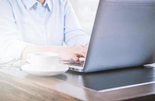 Business concept. Woman in blue shirt typing on computer with coffee on office table, backlighting, sun glare effect, close up, side view, copy space photo