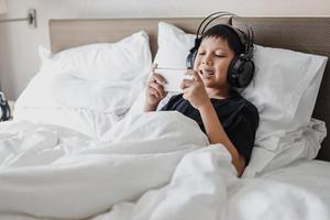 niño asiático feliz con auriculares acostado en la cama mientras juega en un teléfono inteligente