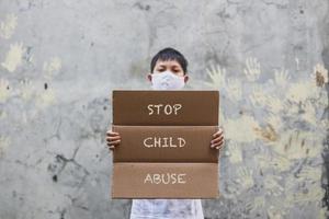 un niño asiático con una máscara médica sosteniendo un tablero de cartas dice que detengan el abuso infantil cuando haga campaña con una huella manual en el fondo de la pared de cemento foto