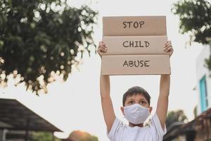 un niño asiático con una máscara médica que levanta un tablero de cartas dice que detengan el abuso infantil cuando haga campaña