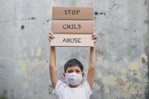 un niño asiático con una máscara médica levantando un tablero de cartas dice que detengan la campaña de abuso infantil con huellas de manos en el fondo de la pared de cemento foto