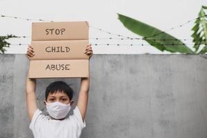 un niño asiático está haciendo campaña con una máscara protectora mientras levanta un tablero de cartas y dice que detengan el abuso infantil foto