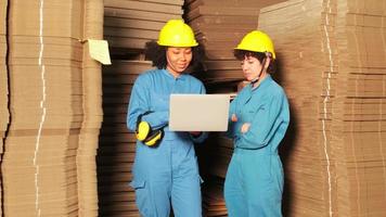 Two female workers and colleagues in safety uniforms and helmet stock check, control production in storage of warehouse factory with pile of stacking cardboard, partner works in manufacture business. video