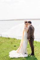 la novia y el novio se abrazan cerca del estanque en la naturaleza. el viento levanta un ligero velo. foto
