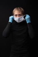 A man in black clothes on a black background in medical gloves puts on a medical mask photo