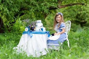 una niña linda con el disfraz de alicia del país de las maravillas organiza una fiesta de té en su mesa mágica. fotografiado en la naturaleza. foto