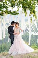 la novia y el novio se abrazan bajo un viejo roble. sesión de fotos de boda en las montañas. junto a ellos se prepara la decoración para la ceremonia.