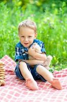 A little cute boy lies on a plaid, hugs his teddy bear. The concept of family holidays in nature.