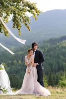 la novia y el novio se abrazan bajo un viejo roble. sesión de fotos de boda en las montañas. junto a ellos se prepara la decoración para la ceremonia.