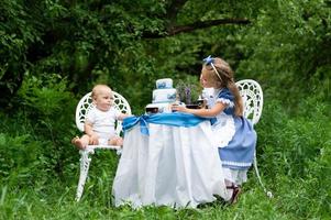 A little cute girl in the costume Alice from Wonderland and her one-year-old brother are having a tea party at their magic table. Photographed in nature. photo