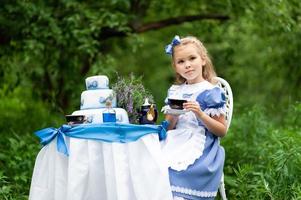 A little cute girl in the costume Alice from Wonderland holds a tea party at her magic table. Photographed in nature. photo