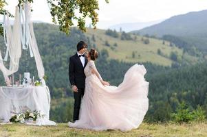 The bride and groom are hugging under an old oak tree. Wedding photo shoot in the mountains. Next to them is prepared decor for the ceremony.