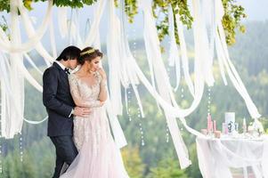 la novia y el novio se abrazan bajo un viejo roble. sesión de fotos de boda en las montañas. junto a ellos se prepara la decoración para la ceremonia.