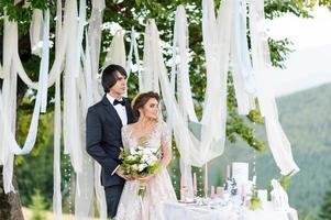 The bride and groom are hugging under an old oak tree. Wedding photo shoot in the mountains. Next to them is prepared decor for the ceremony.