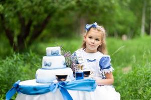 A little cute girl in the costume Alice from Wonderland holds a tea party at her magic table. Photographed in nature.