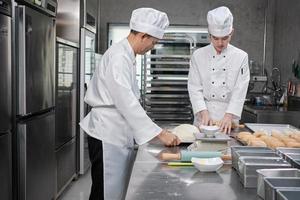 Two professional Asian male chefs in white cook uniforms and aprons are kneading pastry dough and eggs, preparing bread and fresh bakery food, baking in oven at stainless steel kitchen of restaurant. photo