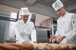 Two professional Asian male chefs in white cook uniforms and aprons are kneading pastry dough and eggs, preparing bread and fresh bakery food, baking in oven at stainless steel kitchen of restaurant.