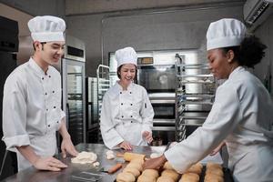 equipo gourmet profesional, tres jóvenes chefs con uniformes de cocina blancos y delantales amasan masa de pastelería y huevos, preparan pan y comida fresca de panadería, horneando en el horno en la cocina del restaurante de acero inoxidable. foto