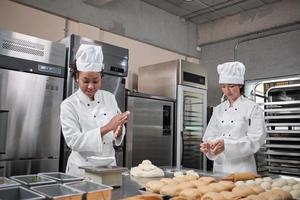 dos chefs profesionales con uniformes y delantales de cocina blanca amasan masa de pastelería y huevos, preparan pan, galletas y comida fresca de panadería, horneando en horno en una cocina de acero inoxidable de un restaurante. foto