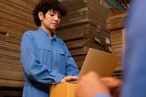 Female delivery worker in safety uniform with a laptop, checks cardboard box, shipping orders at parcels warehouse, paper manufacture factory for the packaging industry, logistic transport service.