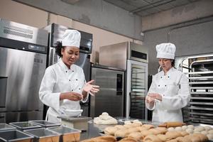 Two professional female chefs in white cook uniforms and aprons knead pastry dough and eggs, prepare bread, cookies, and fresh bakery food, baking in oven at a stainless steel kitchen of a restaurant. photo