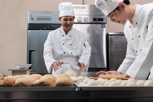 dos chefs asiáticos profesionales con uniformes y delantales de cocinero blanco están amasando masa de pastelería y huevos, preparando pan y comida fresca de panadería, horneando en el horno en la cocina de acero inoxidable del restaurante. foto