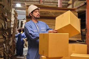 Asian male delivery worker in safety uniform and hard hat checks cardboard box, shipping orders at parcels warehouse, paper manufacture factory for the packaging industry, logistic transport service. photo