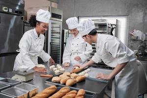 Professional gourmet team, three young chefs in white cook uniforms and aprons knead pastry dough and eggs, prepare bread, and fresh bakery food, baking in oven at stainless steel restaurant kitchen. photo
