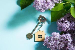 House key with keychain on blue spring background and lilac branches.Summer holiday home, booking cottage in the countryside, moving to new home, mortgage, rent and purchase real estate. Tourism photo