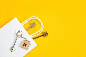 Key ring in the shape of wooden house with key on a yellow background in a white paper gift wrapping bag. Mortgage, rent and purchase real estate, beneficial offer for construction. Copy space photo