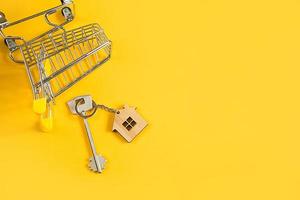 Key ring in the shape of wooden house with key on a yellow background and shopping cart. Mortgage, rent and purchase real estate, spring and summer offer for construction. Copy space photo