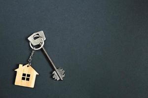 Key chain in the shape of wooden house with key on a black background. Building, design, project, moving to new home, mortgage, rent and purchase real estate. Copy space photo